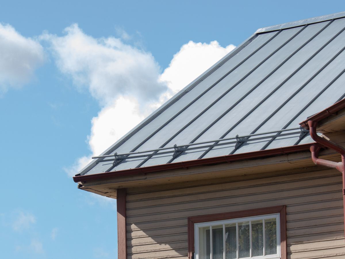 Licensed stock photograph: a pale grey standing seam metal roof on a clapboard house, copper gutter along the eave, blue sky behind.